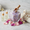 Glass jar filled with colorful pink purple and white bath crystals with wooden scoop surrounded by flower petals on marble surface