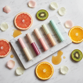 Collection of five elegant lip balm tubes in pastel colors on white marble with rose petals and fresh fruit slices
